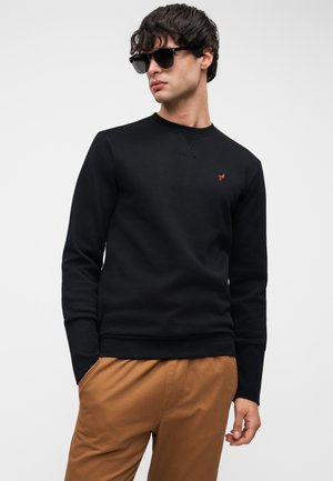 Svart genser med rund hals, teksturert stoff, med en liten rød logo på venstre bryst. Kombinert med brune bukser.