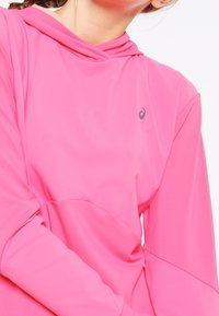Růžová mikina s kapucí vyrobená z měkkého materiálu. Obsahuje zakřivený výstřih a malé logo na hrudi. Dlouhé rukávy a volný střih.