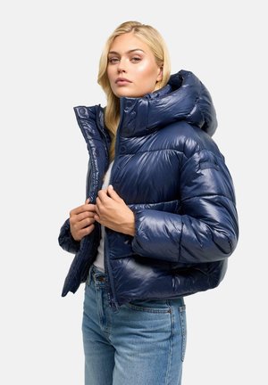 Blondynka ubrana w błyszczącą granatową puchową kurtkę z kapturem, częściowo zapinaną na zamek, w połączeniu z jasnoniebieskimi jeansami, stojąca na białym tle.