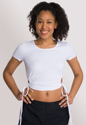 Top corto bianco a coste con maniche corte, lacci laterali per una vestibilità regolabile e scollo rotondo, abbinato a pantaloni scuri e larghi.
