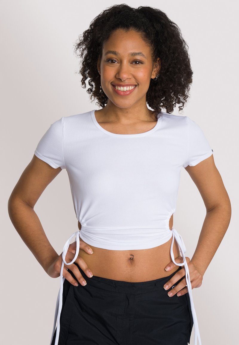 Top corto bianco a coste con maniche corte, lacci laterali per una vestibilità regolabile e scollo rotondo, abbinato a pantaloni scuri e larghi.