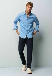 Lichtblauw denim overhemd met lange mouwen, knoopsluiting aan de voorkant en borstzak, gecombineerd met donkere broeken en crèmekleurige sneakers.