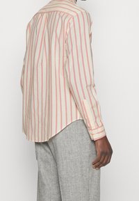 Camisa a rayas en color crema con líneas verticales rojas, de mangas largas y con un dobladillo curvado. El material parece ligero y tiene una textura suave.