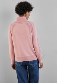 Rosa Pullover-Sweater mit hohem Kragen, gerippten Ärmelbündchen und Saum. Verfügt über ein strukturiertes Muster auf den Schultern. Getragen mit blauen Jeans.