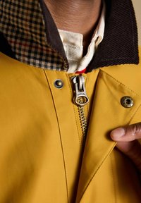 Close-up van een persoon die een mosterdgele jas draagt met een geruite kraag en een deels dichtgezette metalen rits met het label "Joules".