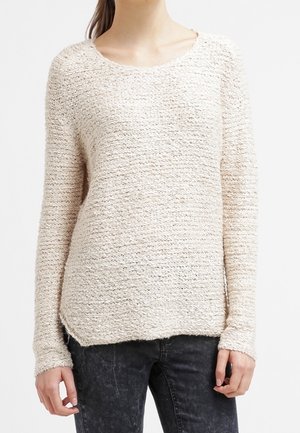 Femme portant un pull en maille texturé crème à manches longues et col rond, associé à un jean noir délavé.