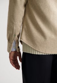 Beige uldblandingsskjorte med synlig tekstur, sideslids og ribbet grøn kant. Har knapper i grøn og sort samt stribede skjortearmer.