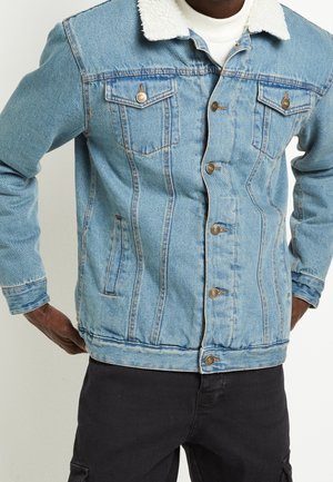 Hombre con chaqueta vaquera azul claro con cuello de borrego blanco y pantalones negros, con las manos apoyadas en las caderas.