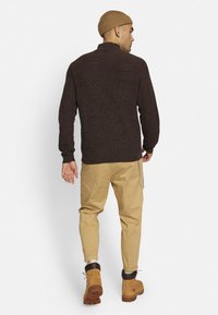 Brun ribbet sweater med høj krave, båret med lysebrune tætsiddende bukser og brune støvler, der har kædeddetaljer på bukserne.