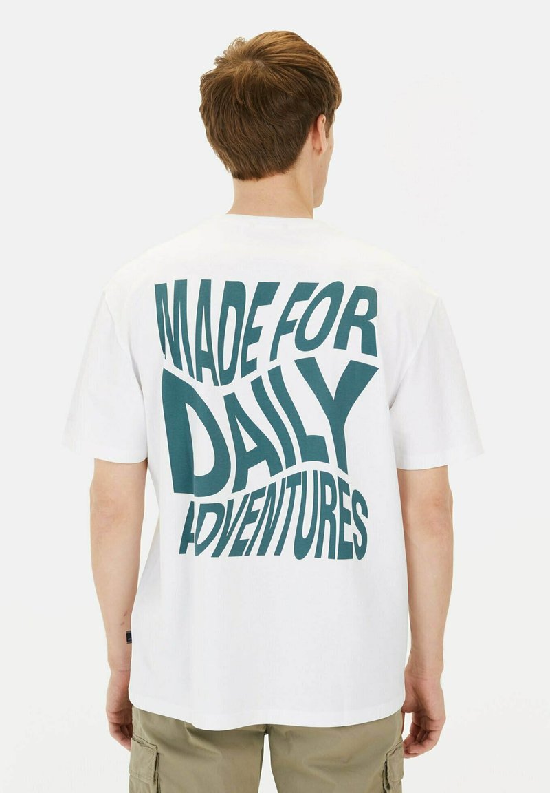 Witte katoenen t-shirt met een gedurfde, gebogen donkerblauwe tekstontwerp op de achterkant, met de tekst "MADE FOR DAILY ADVENTURES," korte mouwen, losse pasvorm.