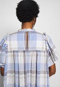 Osoba s krátkými kudrnatými vlasy nosí rýhované modro-bílé kostkované šaty s výstřihem na zádech a volánkovými rukávy.