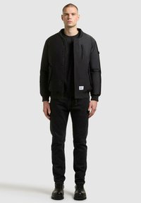 Schwarze Bomberjacke mit Reißverschlusstasche, Druckknopfverschluss und gerippten Bündchen. Kombiniert mit schwarzer Jeans und robusten schwarzen Stiefeln.