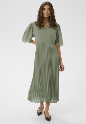 Kobieta o kręconych włosach, ubrana w plisowaną sukienkę midi w kolorze szałwiowym z bufkami na rękawach i czarne, szpiczaste sandały na obcasie ze zapięciem z tyłu, stojąca na białym tle.