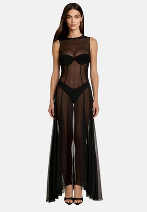 Donna con lunghi capelli scuri che indossa un abito nero trasparente senza maniche sopra un reggiseno e biancheria nera, in piedi davanti a uno sfondo chiaro e semplice.