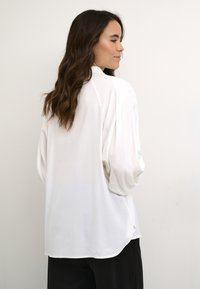 Vit långärmad blus med avslappnad passform, puffärmar och en mjuk textur. Har en hög halsringning och en rundad fåll.
