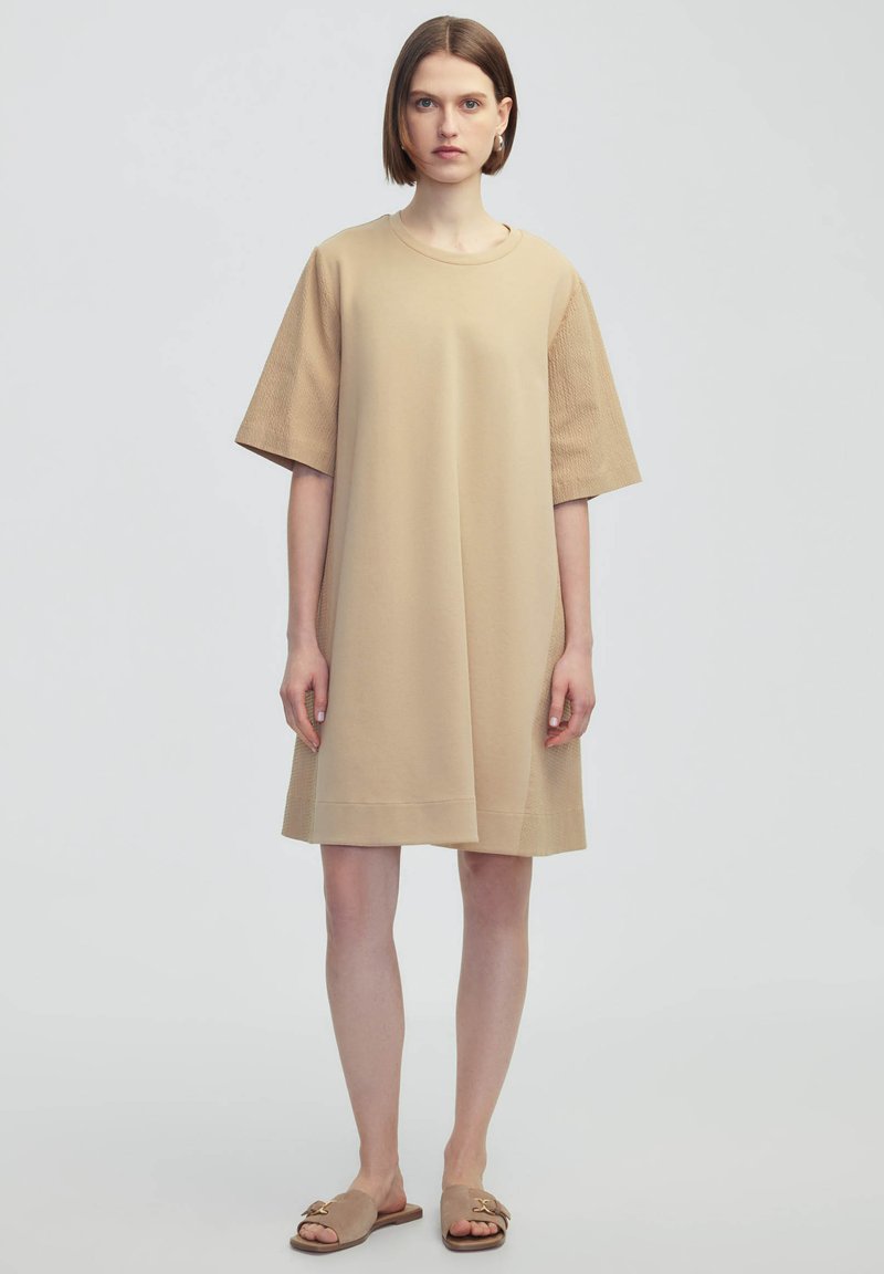 Vestido beige de corte oversize con mangas cortas texturizadas, cuello redondo y corte recto. Llevado con sandalias de slide en marrón claro.