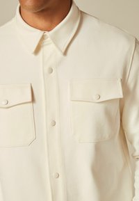 Lys beige button-up skjorte i tekstureret stof med to brystlommer og spids krave. Disciple knapper og syninger.