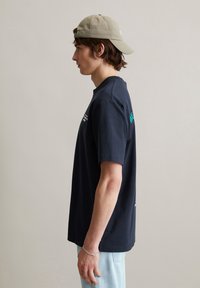 Marineblauwe katoenen T-shirt met korte mouwen, ronde hals en logo-accenten op de voor- en achterkant, gecombineerd met lichtblauwe jeans en een beige pet.