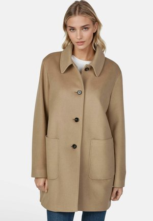Femme portant un manteau en laine beige avec un grand col, des boutons noirs et deux poches avant, par-dessus un jean bleu et une chemise blanche.