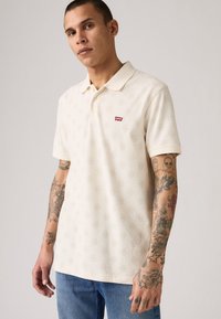 Cremfarbene Polo-Shirt mit strukturiertem Muster, geripptem Kragen und kleinem roten Levi's-Logo auf der Brust. Kurze Ärmel und lockere Passform.