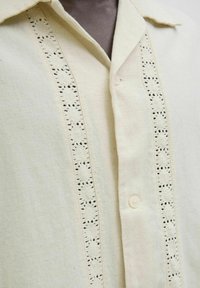 Camisa color crema con cierre de botones en la parte delantera, que cuenta con un borde de encaje en el centro y un cuello puntiagudo. Textura suave, tejido ligero.