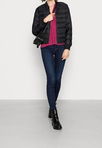 Doudoune noire avec un ourlet côtelé, haut violet, jeans foncés et bottines noires à accents de boucle. Modèle portant un sac à main noir.