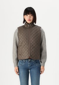 Chaleco acolchado verde oliva con cuello alto, cierre de cremallera y sin mangas. Lleva puesto sobre un suéter gris con puños acanalados, combinado con jeans azules.