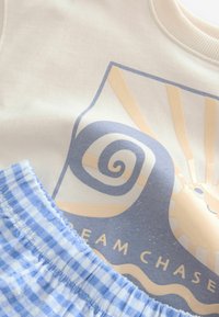 Felpa crema con design grafico blu che presenta una spirale e un sole, abbinata a pantaloni corti a quadretti blu chiaro e bianchi con vita elastica.