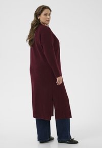 Cardigan lungo borgogna con texture a coste, spacchi laterali e vestibilità ampia. Abbinato a jeans svasati scuri e scarpe basse nere.