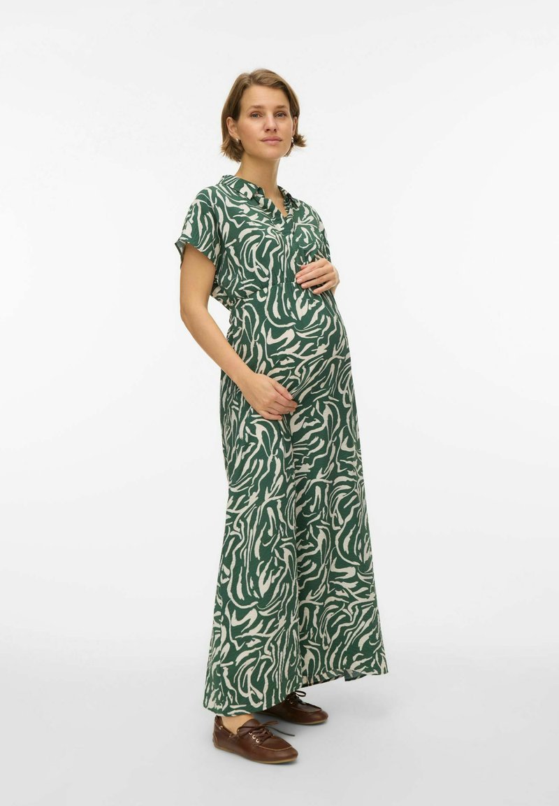 Femme enceinte en robe maxi à motif vert et blanc, tenant son ventre, debout devant un fond blanc uni.