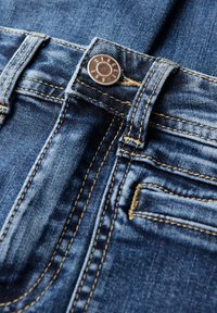 Jeans in denim di colore blu scuro, con hardware in bottone di rame e cuciture gialle a contrasto. Include un piccolo dettaglio della tasca frontale.