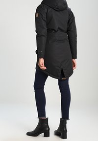 Czarna ocieplana kurtka z kapturem, regulowanym pasem i naszywką z logo. Noszona na ciemnych dżinsach i czarnych botkach za kostkę o teksturowanym wykończeniu.