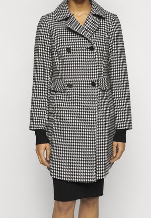Femme portant un manteau croisé pied-de-poule noir et blanc avec des boutons noirs sur une jupe noire sur un fond uni.