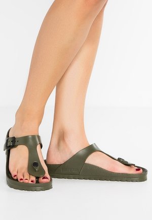 Stopy w oliwkowozielonych sandałach Birkenstock z regulowanymi klamrami, ukazujące pomalowane na ciemnoczerwony kolor paznokcie na białym tle.