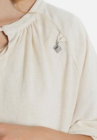 Beige top met korte mouwen en een geplooide halslijn. Heeft een klein zilveren label of bedel dat de textuur en eenvoud benadrukt.