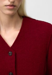 Czerwony, teksturowany kardigan z dekoltem w kształcie V, z czarnymi guzikami z przodu i ciepłym, miękkim materiałem. Zbliżenie koncentruje się na tkaninie i detalach.