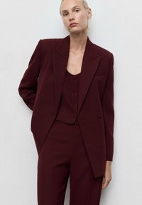 Burgundin räätälöity bleiseri terävillä läpillä, yhdistettynä samasta kankaasta valmistettuun hihattomaan toppiin, jossa on kolme nappia, sekä slim fit -housuihin.