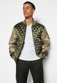 Veste bomber matelassée en vert olive avec des manches dorées métalliques. Comprend un col et des poignets côtelés, une fermeture éclair et des détails de couture.