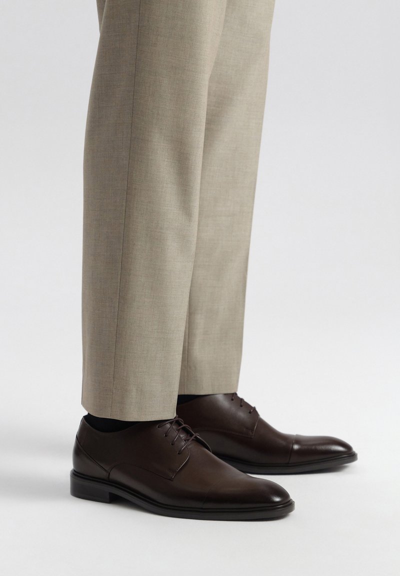 Zapatos de vestir de cuero marrón con un acabado suave, diseño con cordones y punta redonda, combinados con pantalones de vestir beige claros.