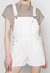 Persoon draagt witte denim korte overalls met rafelige zomen over een taupekleurige top met korte mouwen, zichtbaar vanaf de schouders tot halverwege de dijen.