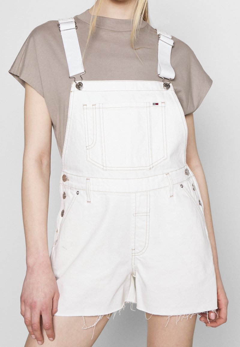 Persoon draagt witte denim korte overalls met rafelige zomen over een taupekleurige top met korte mouwen, zichtbaar vanaf de schouders tot halverwege de dijen.