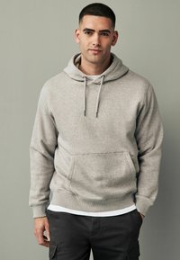 Sudadera gris con un bolsillo frontal, capucha con cordones, textura suave y puños acanalados, llevada sobre una camisa blanca, combinada con pantalones cargo oscuros.