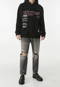 Černá mikina s kapucí s růžovým a bílým textem, doplněná o potrhané šedé džíny. Celkový vzhled dotvářejí černé tenisky.