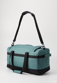 Sac de sport en tissu teal avec des accents noirs, doté de doubles poignées et d'une bandoulière amovible. Texture lisse, forme rectangulaire.