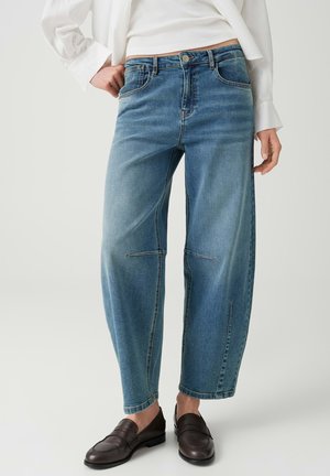 Personne portant un jean ample bleu clair et des mocassins marron foncé, avec une main posée sur la hanche et une chemise blanche visible.