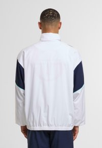 Fehér dzseki tengerészkék díszítéssel, elasztikus derékpánttal, magas gallérral és perforált mintás logórészlettel a háton. Simább anyag.