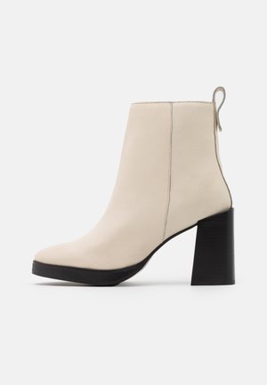 Bottine beige en cuir avec talon haut noir épais et languette à l'arrière, vue de profil sur fond blanc.