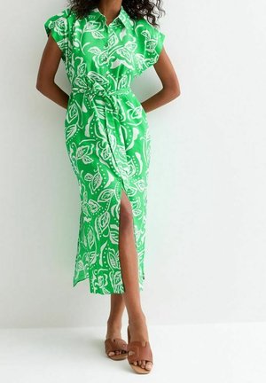 Femme portant une robe midi verte avec des motifs de feuilles blanches, une ceinture à la taille, une fente à l'avant et des sandales marron à bout ouvert.