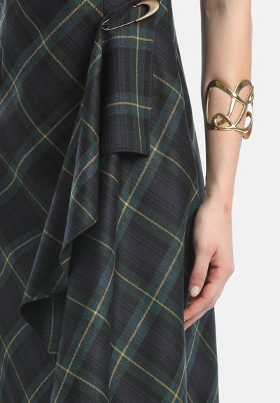 Braccio con bracciale aperto in oro accanto a tessuto a quadri verde scuro e blu navy, fissato con una grande spilla di sicurezza dorata.