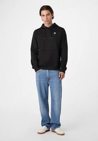 Czarna bluza z kapturem z przednią kieszenią i wyszywanym logo, zestawiona z jasnoniebieskimi dżinsami o szerokich nogawkach oraz białymi trampkami.